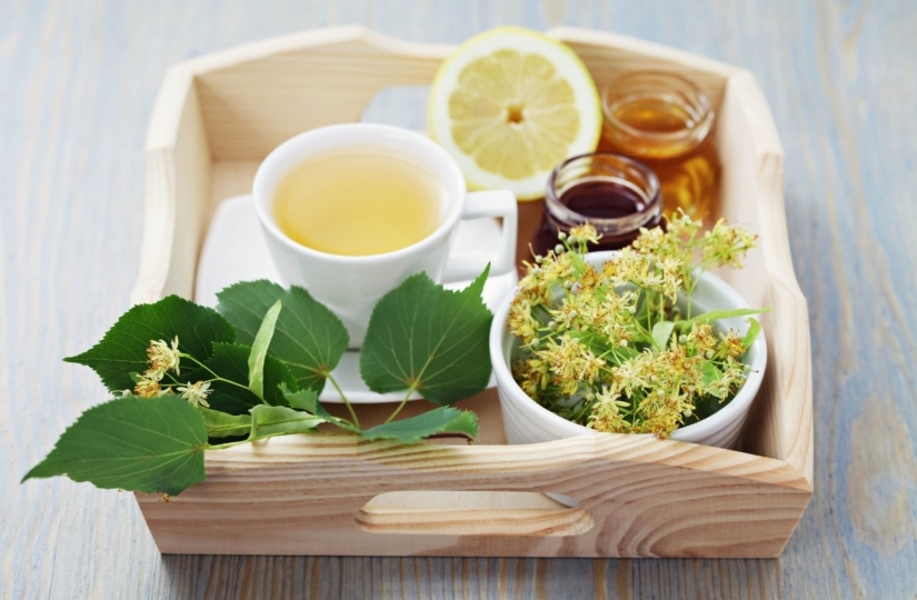 wooden tray holding a mug of green tea, a lemon, a jar of honey and a bowl of wildflowers