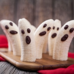 homemade halloween scary banana ghosts monsters with chocolate faces. healthy natural vegetarian snack funny dessert recipe for party decoration on vintage wooden table background and red fabric.