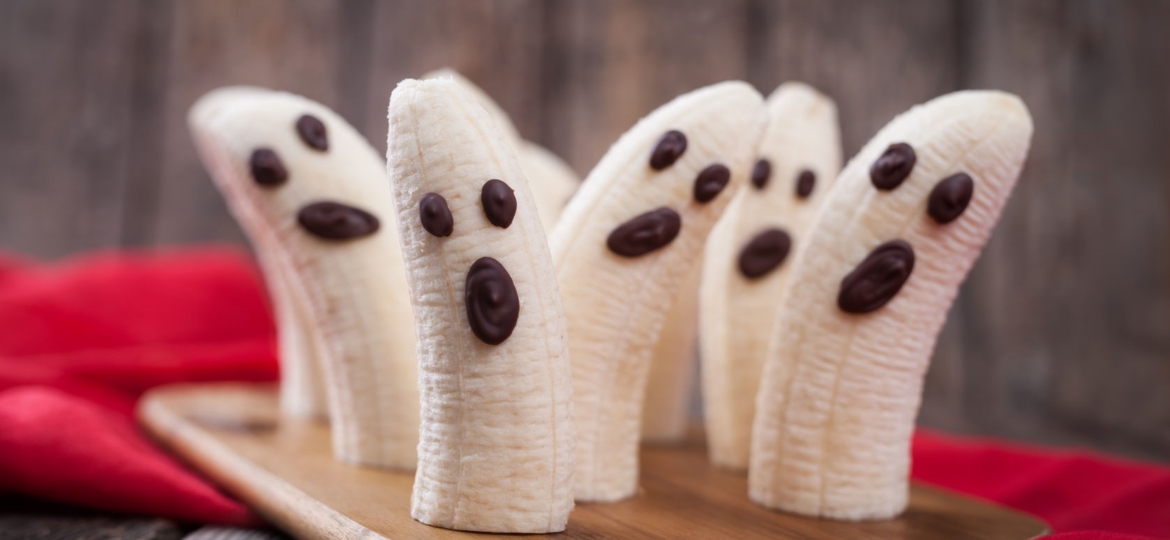 homemade halloween scary banana ghosts monsters with chocolate faces. healthy natural vegetarian snack funny dessert recipe for party decoration on vintage wooden table background and red fabric.